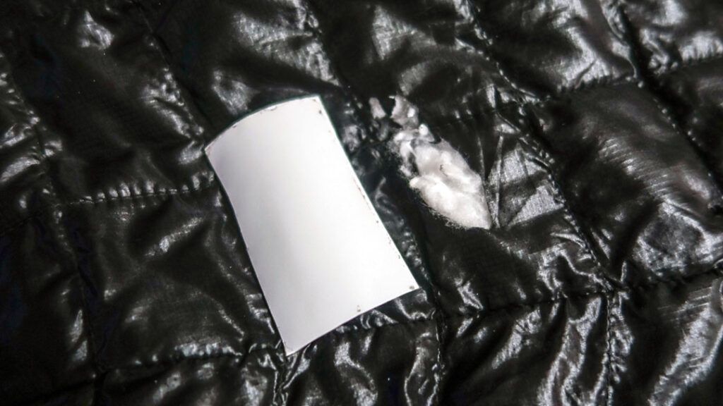 A rectangular patch of Tenacious Tape cut from the roll sits next to a hole in the puffer jacket.