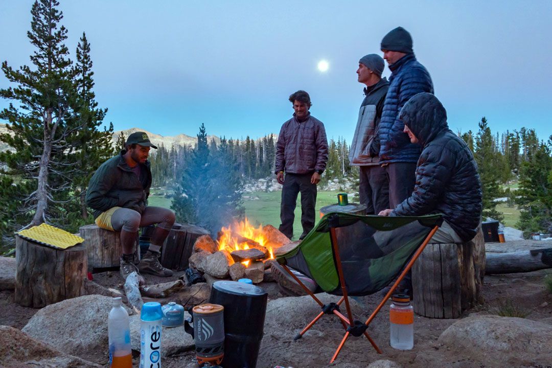 Friends gather around a campfire near a meadow at moonrise.